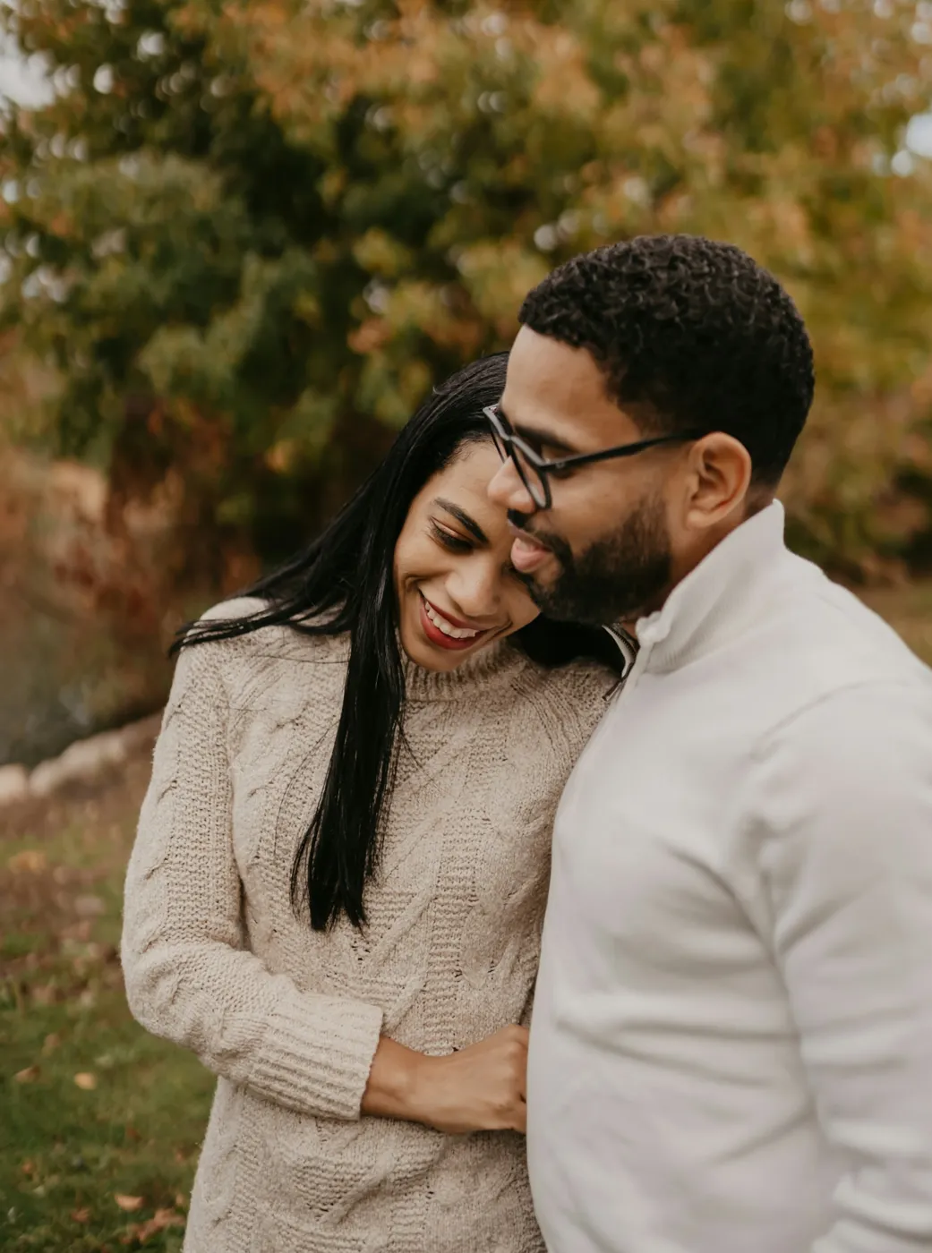 A wife rests her head on her husband's shoulder as they laugh together outdoors in autumn